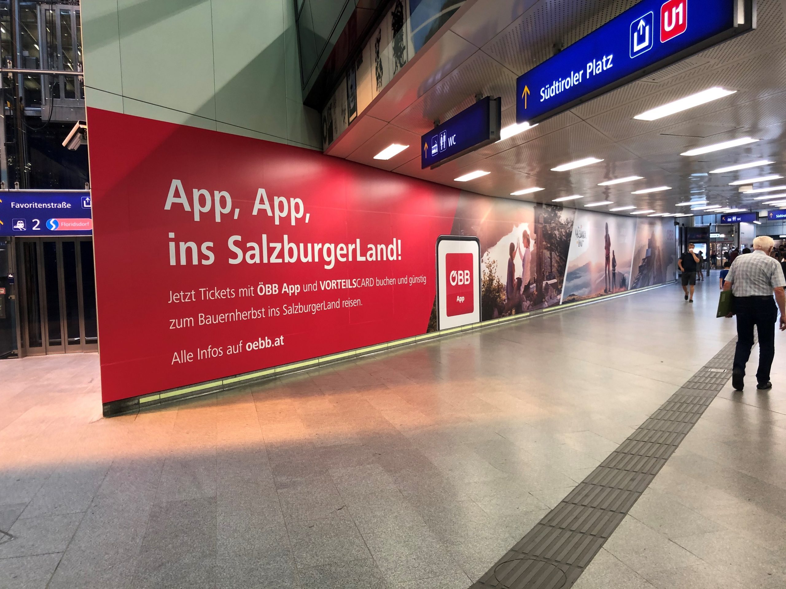 Große rote Werbung in einem Bahnhof, die für die ÖBB-App im SalzburgerLand wirbt, mit vorbeilaufenden Reisenden.