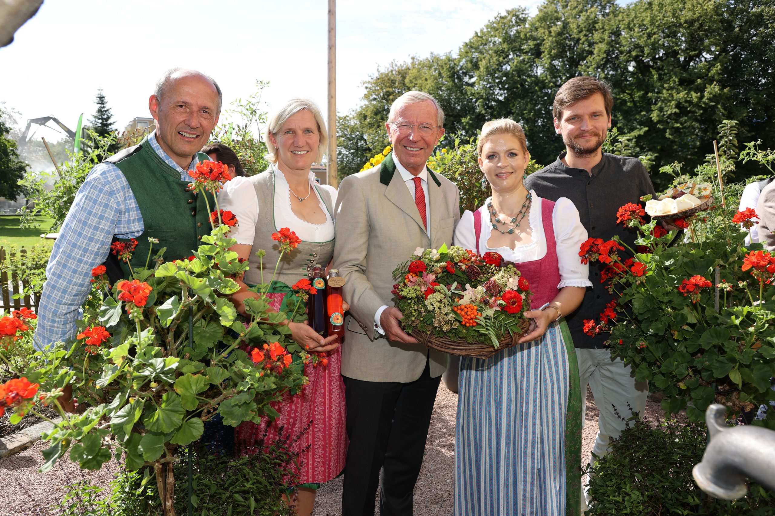 Fünf Personen in österreichischer Tracht stehen im Freien zwischen Blumen, lächeln und halten einen Korb mit Blüten.