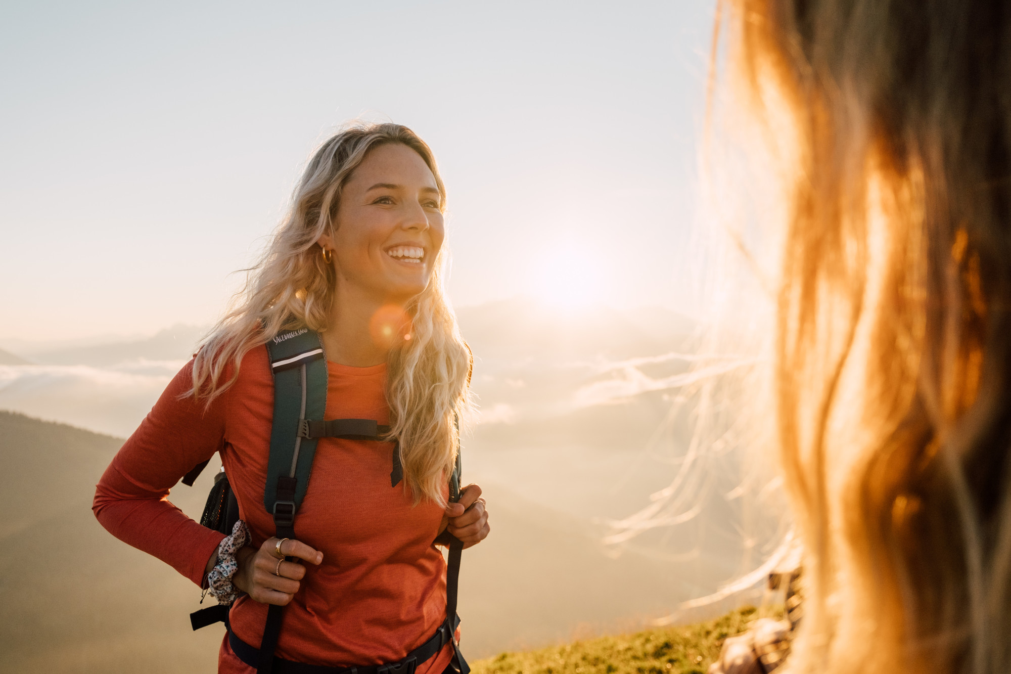 Lächelnde Frau mit einem Rucksack wandert bei Sonnenaufgang, Berge und Sonnenlicht im Hintergrund.