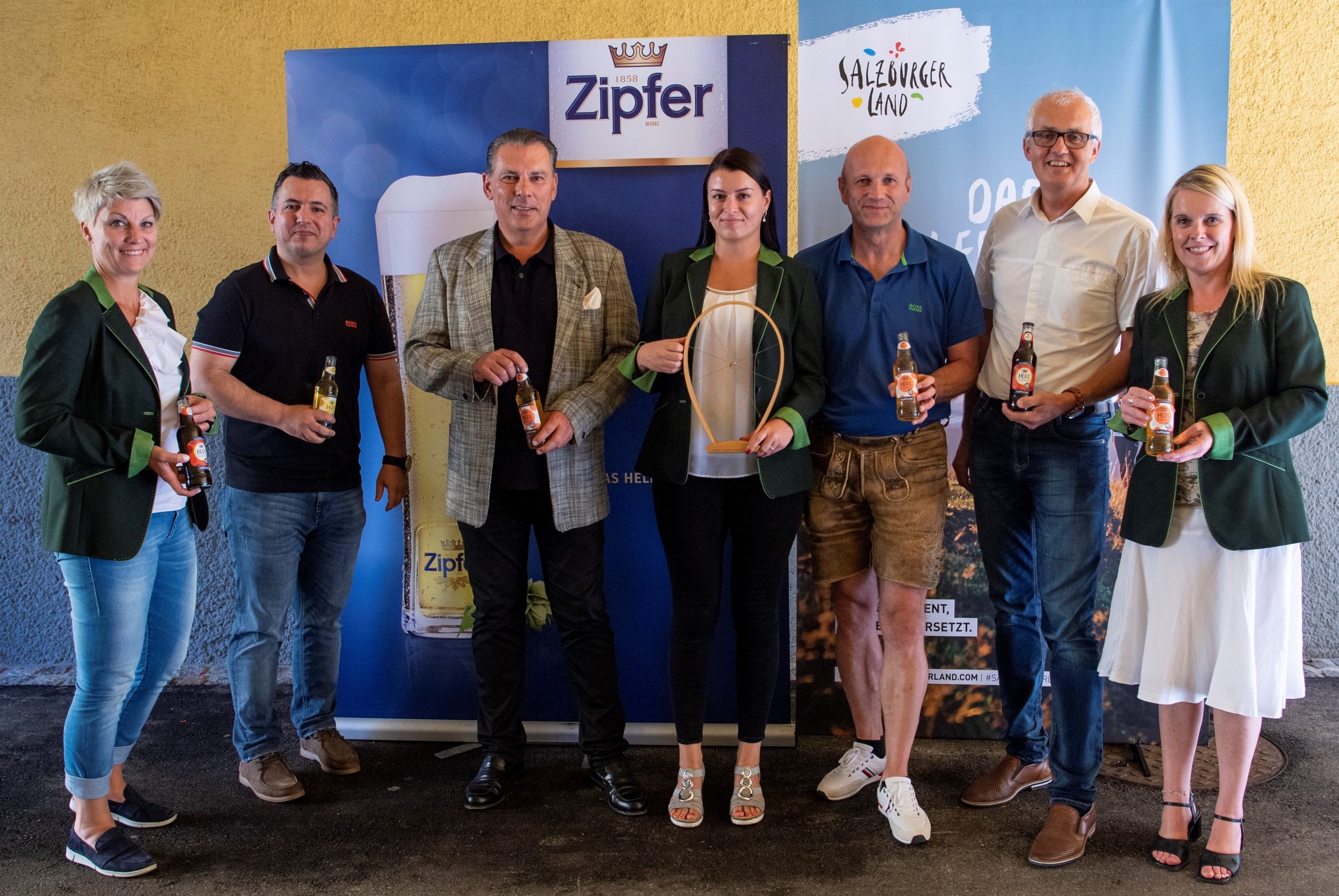 Sieben Personen stehen lächelnd und mit Bierflaschen in der Hand vor Zipfer- und Salzburger Land-Transparenten.