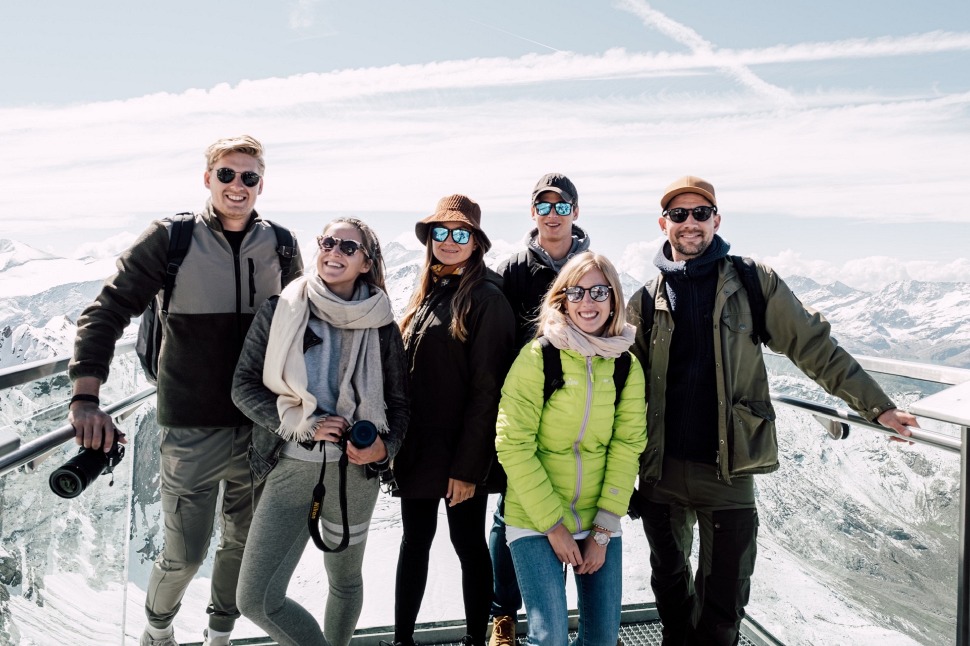 Sechs Personen in Winterkleidung posieren auf einer verschneiten Aussichtsplattform in den Bergen mit blauem Himmel und Wolken im Hintergrund.