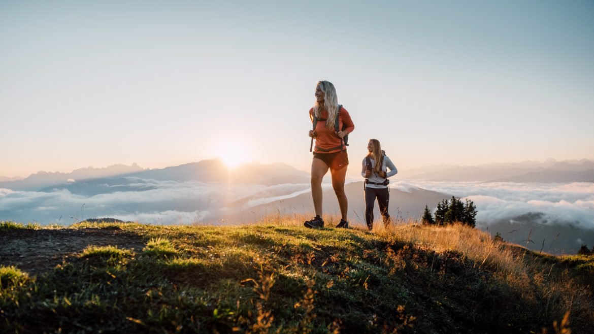 Zwei Frauen wandern bei Sonnenaufgang über einen grasbewachsenen Hügel - Momente wie diese, dafür leben wir.