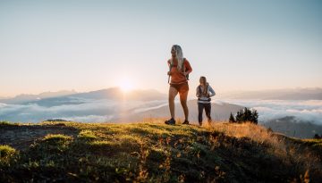 Zwei Frauen wandern bei Sonnenaufgang über einen grasbewachsenen Hügel - Momente wie diese, dafür leben wir.