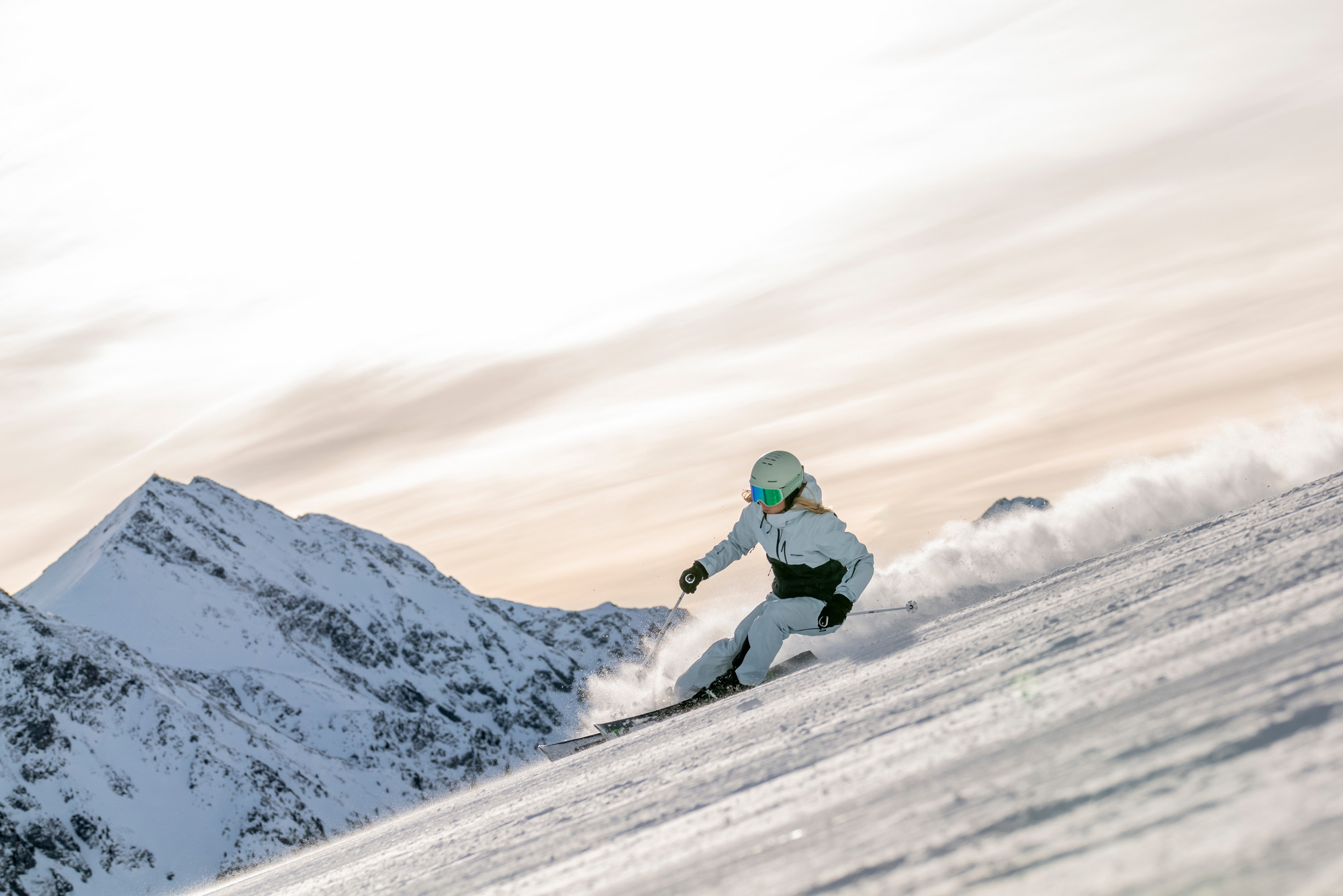 Ein Skifahrer in weißer Kleidung fährt auf einem verschneiten Berghang mit Gipfeln im Hintergrund bergab.
