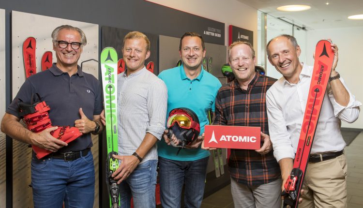 Fünf lächelnde Männer halten Atomic-Ski und -Ausrüstung und heben Wirtschaftskooperationen vor einer Skiwand hervor.