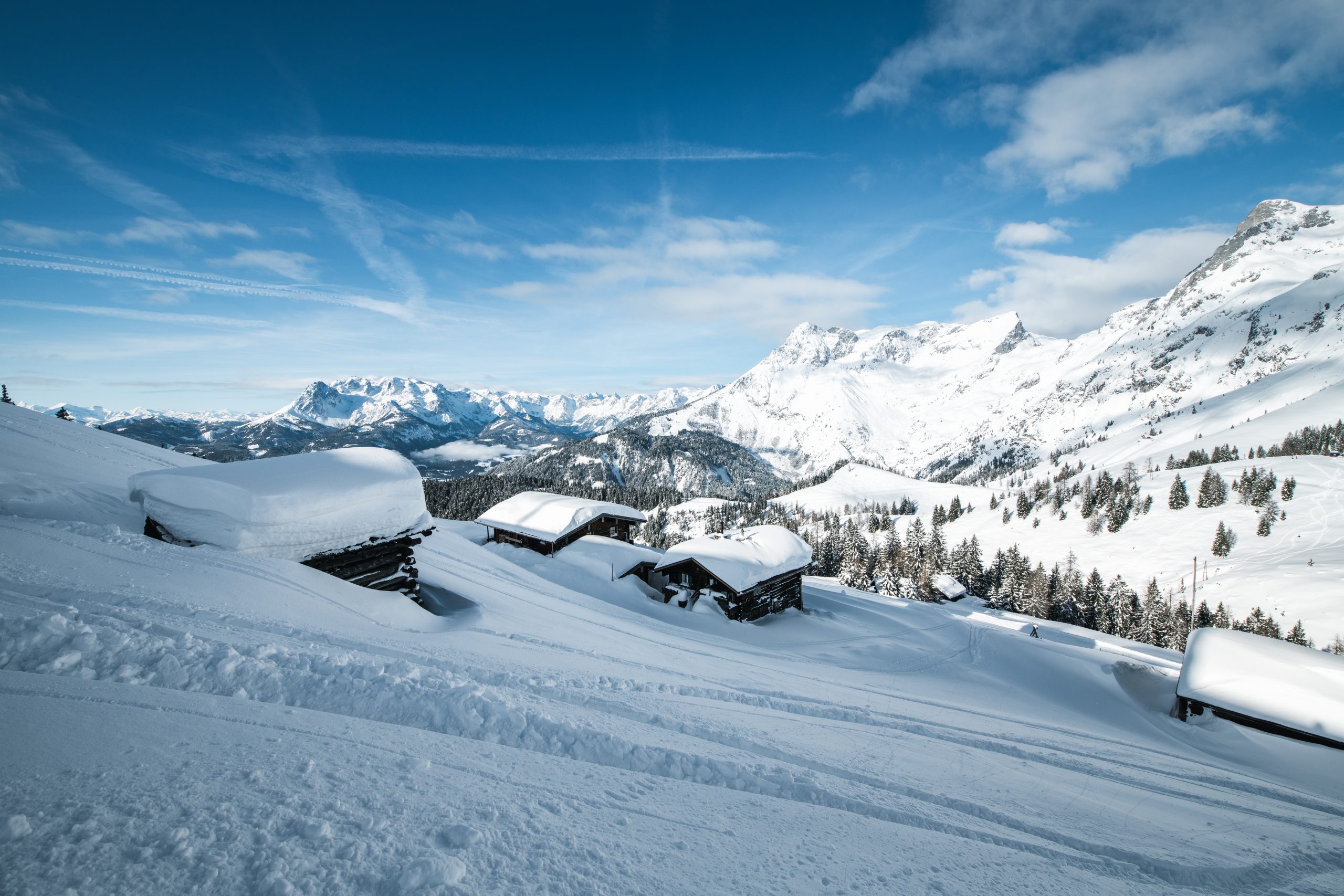 Schneebedeckte Hütten an einem Hang mit hohen Bergen und blauem Himmel im Hintergrund.