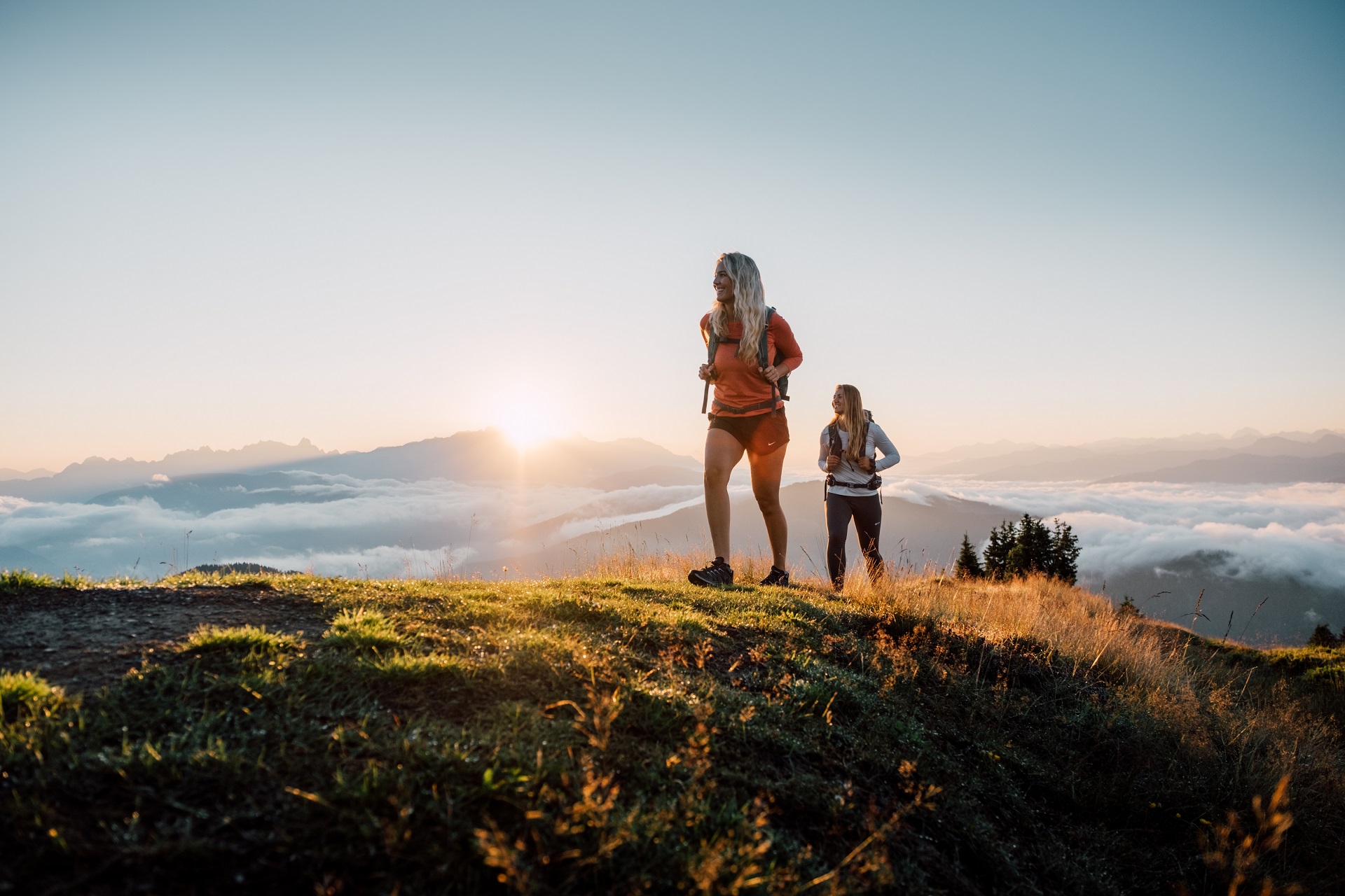 Zwei Frauen wandern auf einem grasbewachsenen Hügel bei Sonnenaufgang, mit Bergen und Wolken im Hintergrund.