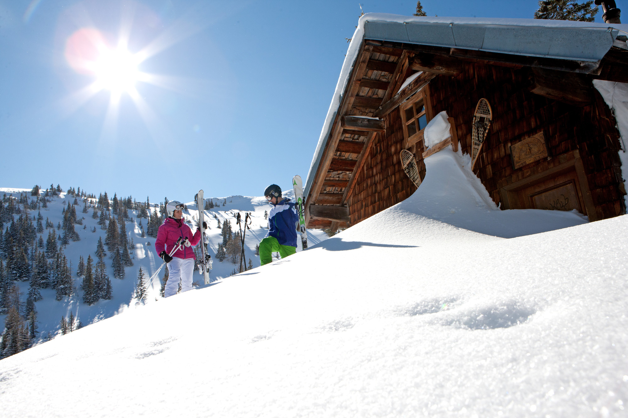 Zwei Skifahrer stehen an einem sonnigen Tag in den Bergen vor einer verschneiten Holzhütte, die Bäume und den blauen Himmel im Blick.