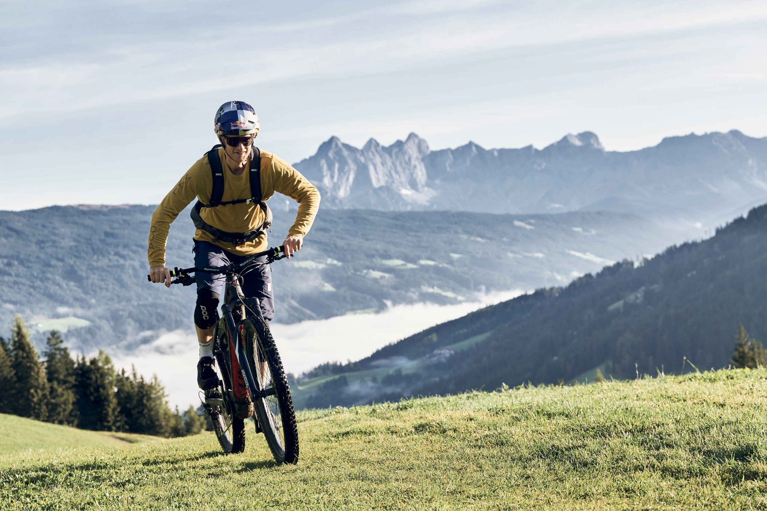 Eine Person fährt mit einem Mountainbike bergauf auf einem grasbewachsenen Hang mit Bergen im Hintergrund.