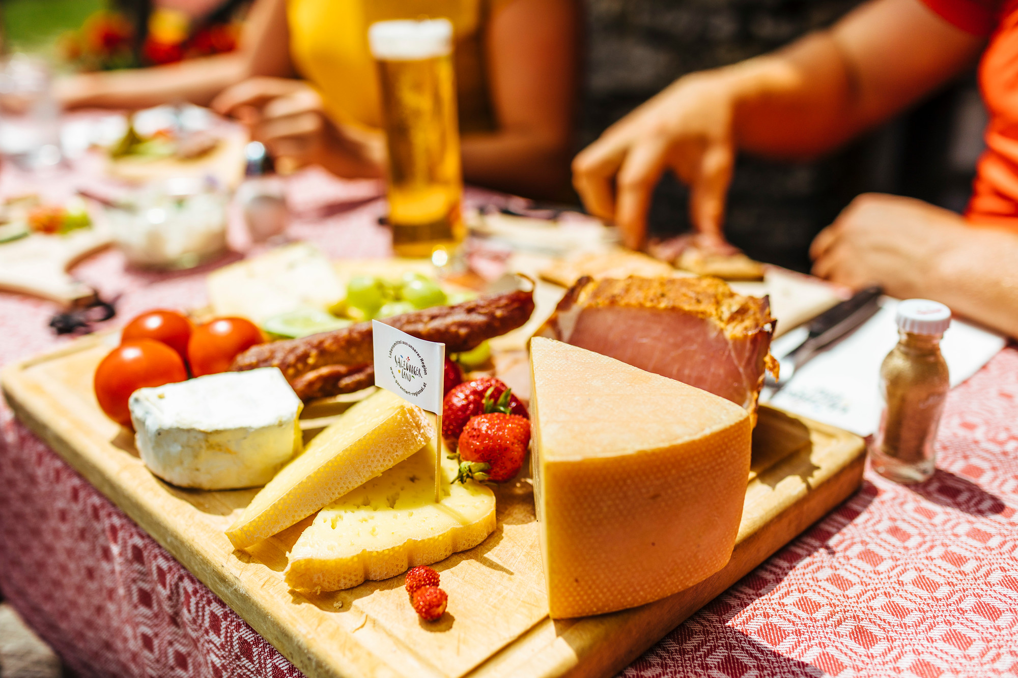 Ein Holzbrett mit verschiedenen Käsesorten, Wurstwaren, Tomaten und Erdbeeren auf einem Tisch im Freien.