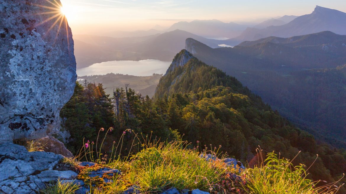 Berglandschaft bei Sonnenaufgang mit Sonnenaufgang, See und üppigen grünen bewaldeten Hügeln in der Ferne.
