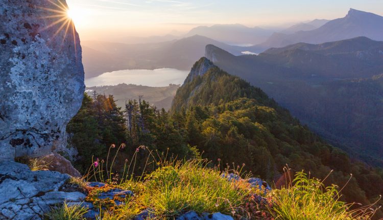 Berglandschaft bei Sonnenaufgang mit Sonnenaufgang, See und üppigen grünen bewaldeten Hügeln in der Ferne.