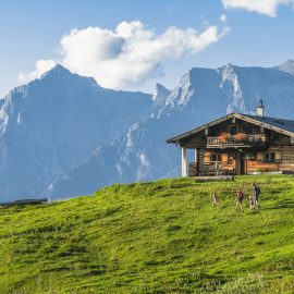 Holzhütte auf einem grünen Hügel mit spazierenden Menschen vor hohen, felsigen Bergen und einem blauen Himmel.