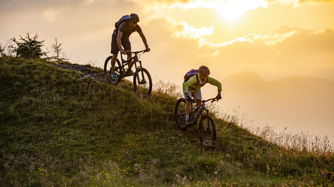Zwei Radfahrer fahren mit ihren Mountainbikes bergab auf einem grasbewachsenen Weg bei Sonnenuntergang, während der Himmel im Hintergrund leuchtet.