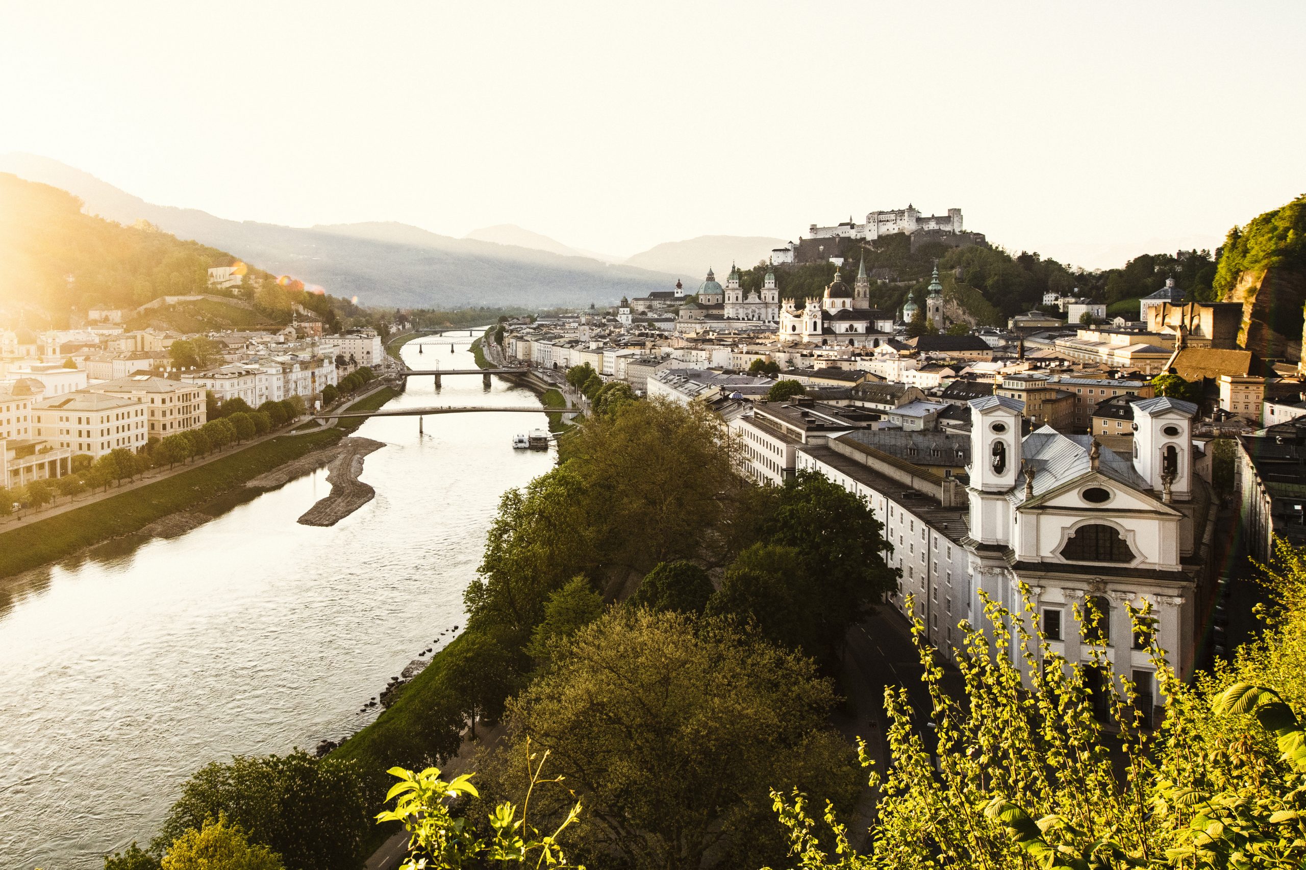 Eine malerische Ansicht von Salzburg, Österreich, mit einem Fluss, Brücken und historischen Gebäuden bei Sonnenuntergang.