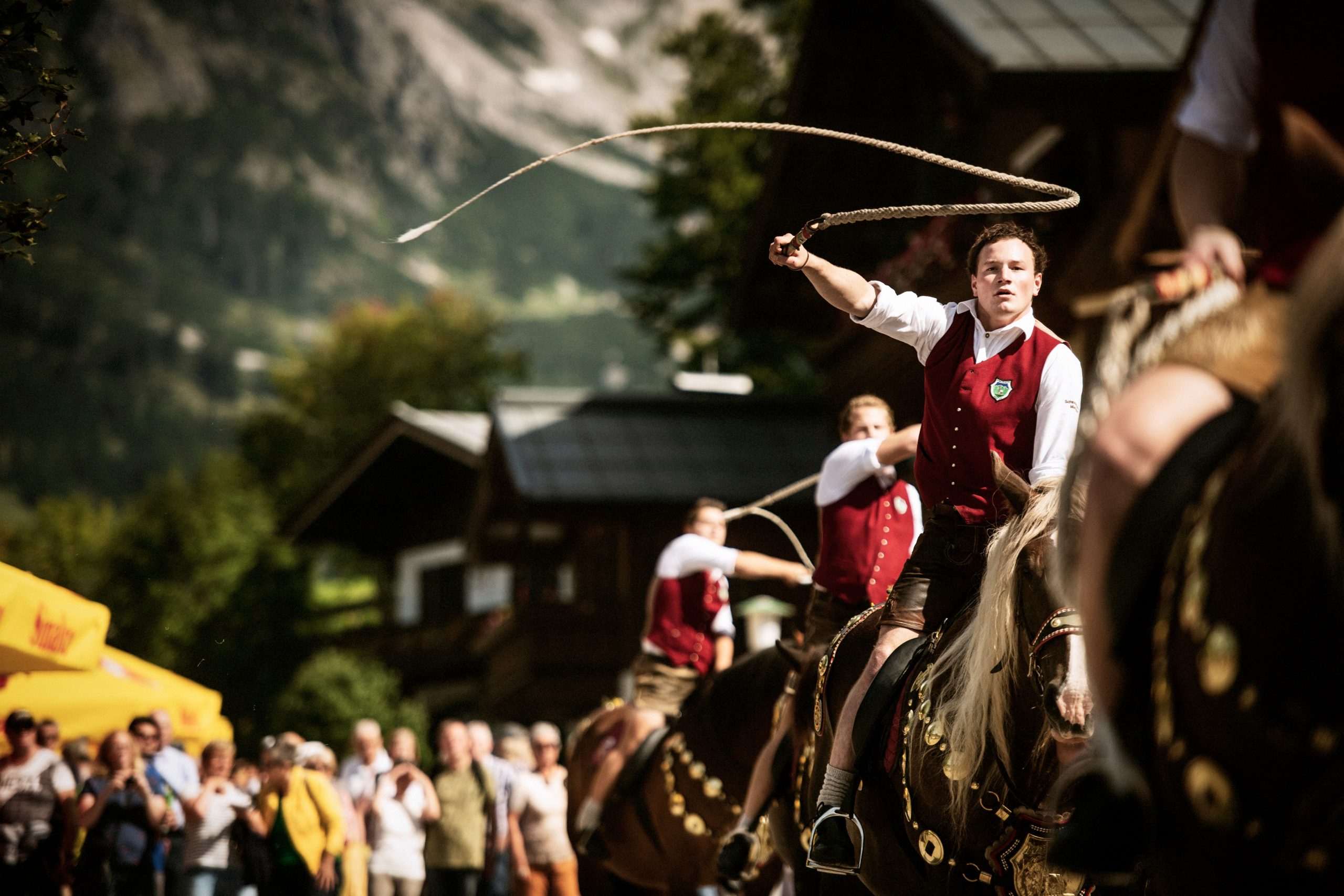 Ein Mann in traditioneller österreichischer Kleidung auf einem Pferd knallt die Peitsche während eines Festes mit einer Menschenmenge und Bergen im Hintergrund.