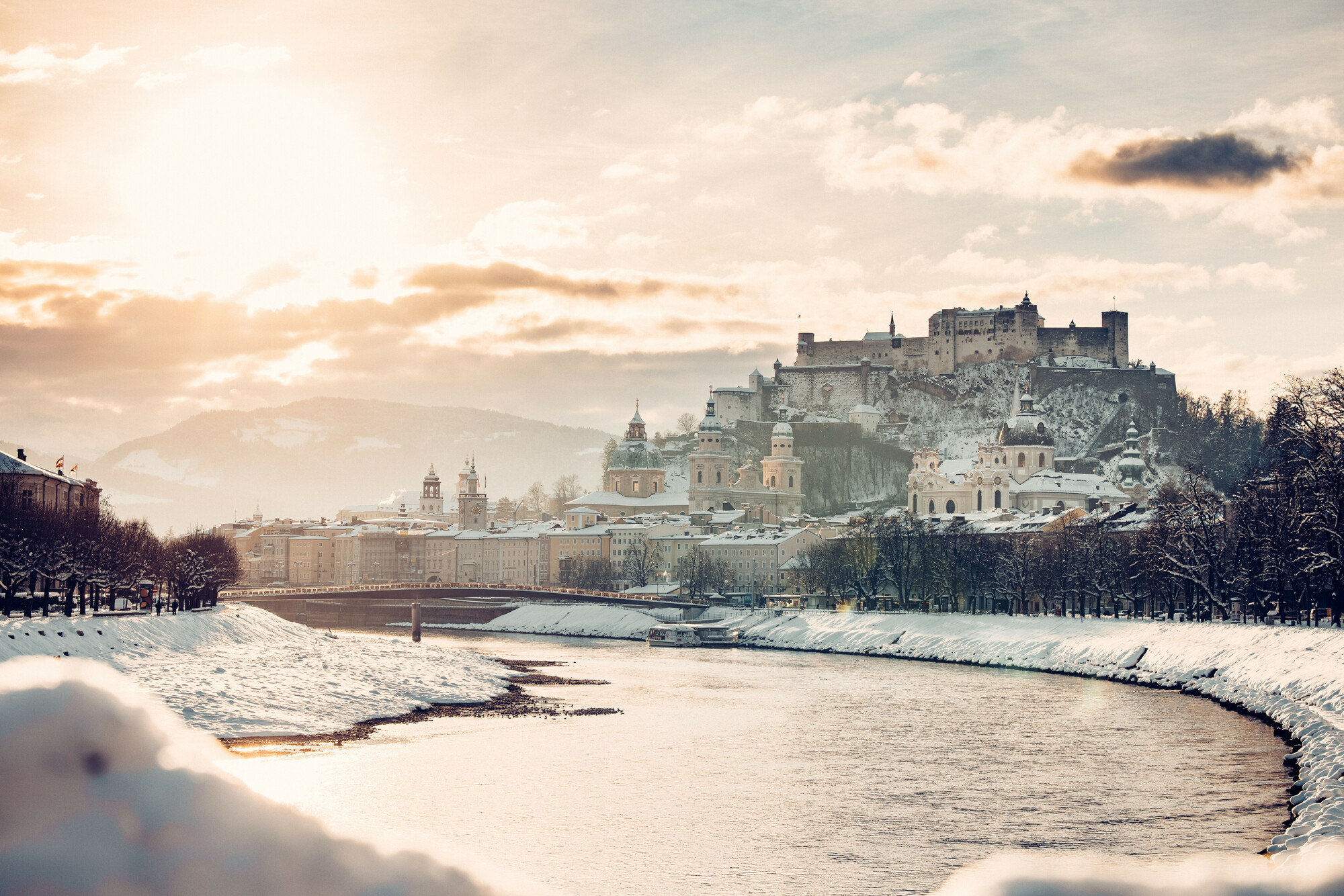Verschneites Stadtbild von Salzburg mit einem Fluss, Kuppelkirchen und einer Festung auf dem Hügel bei Sonnenuntergang.