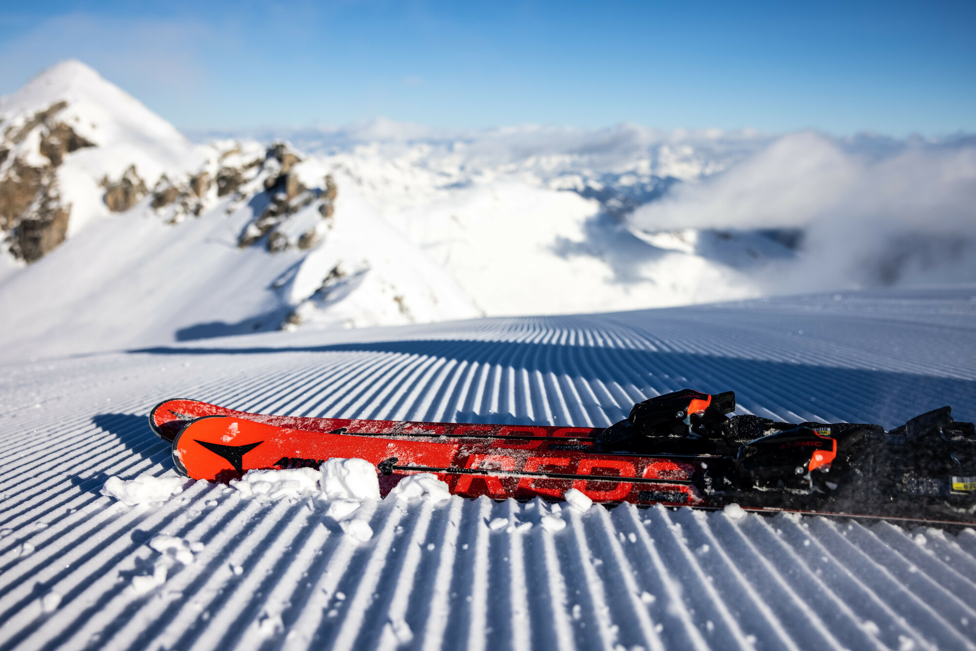Ein Paar rote Skier, die auf frisch präpariertem Schnee ruhen, mit verschneiten Bergen im Hintergrund.