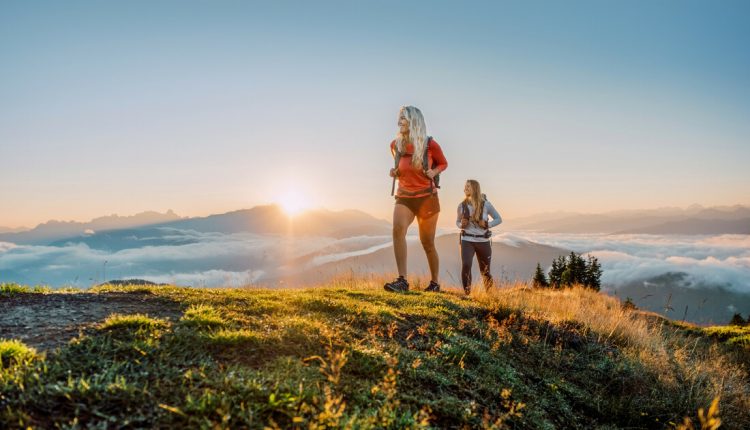 Zwei Frauen wandern bei Sonnenaufgang über einen grasbewachsenen Hügel mit den Bergen der SalzburgerLand Tourismus GmbH im Hintergrund.