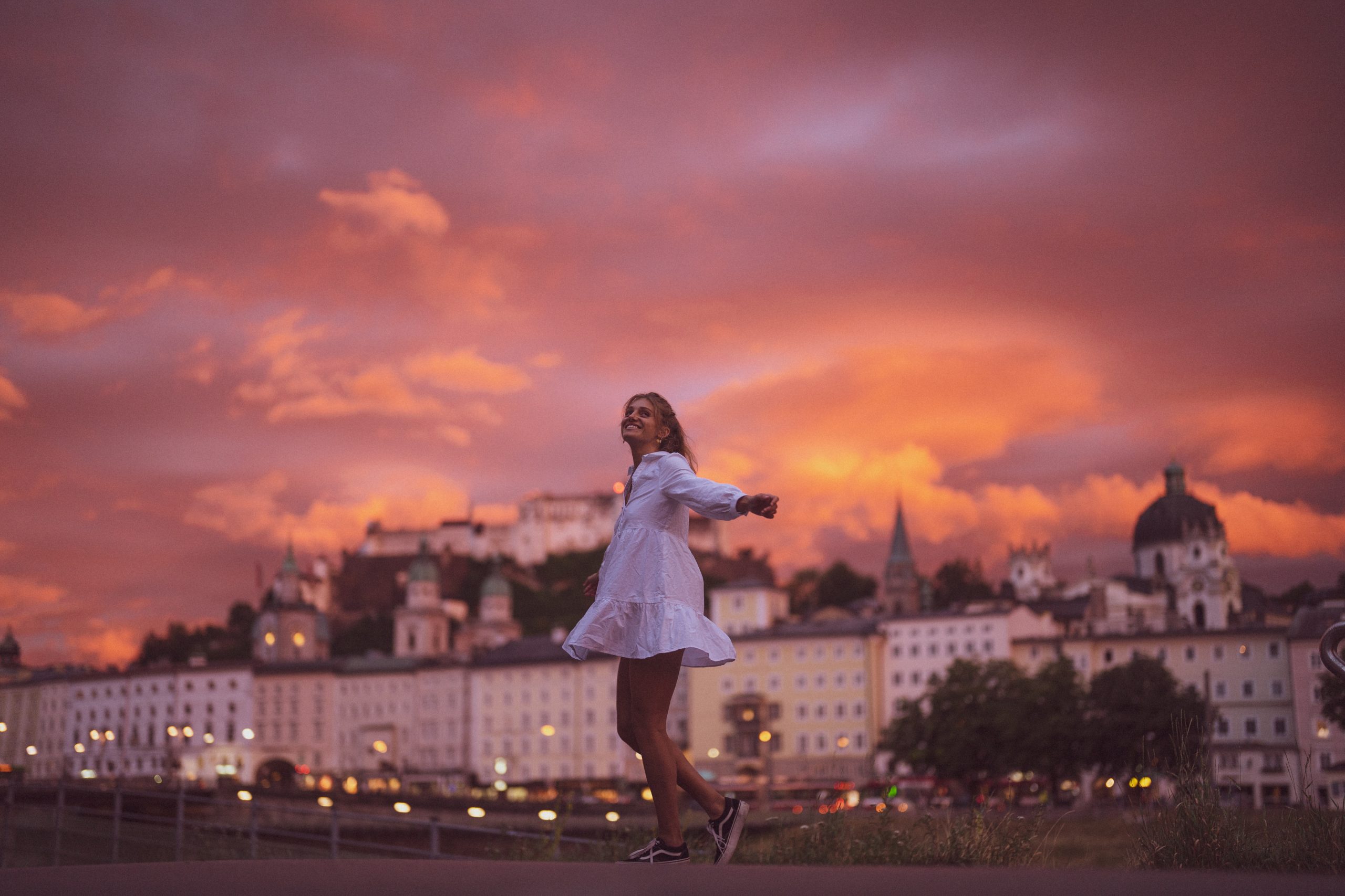Eine Frau in einem weißen Kleid wirbelt fröhlich bei Sonnenuntergang, mit einer Stadt und einem dramatischen orangefarbenen Himmel im Hintergrund.