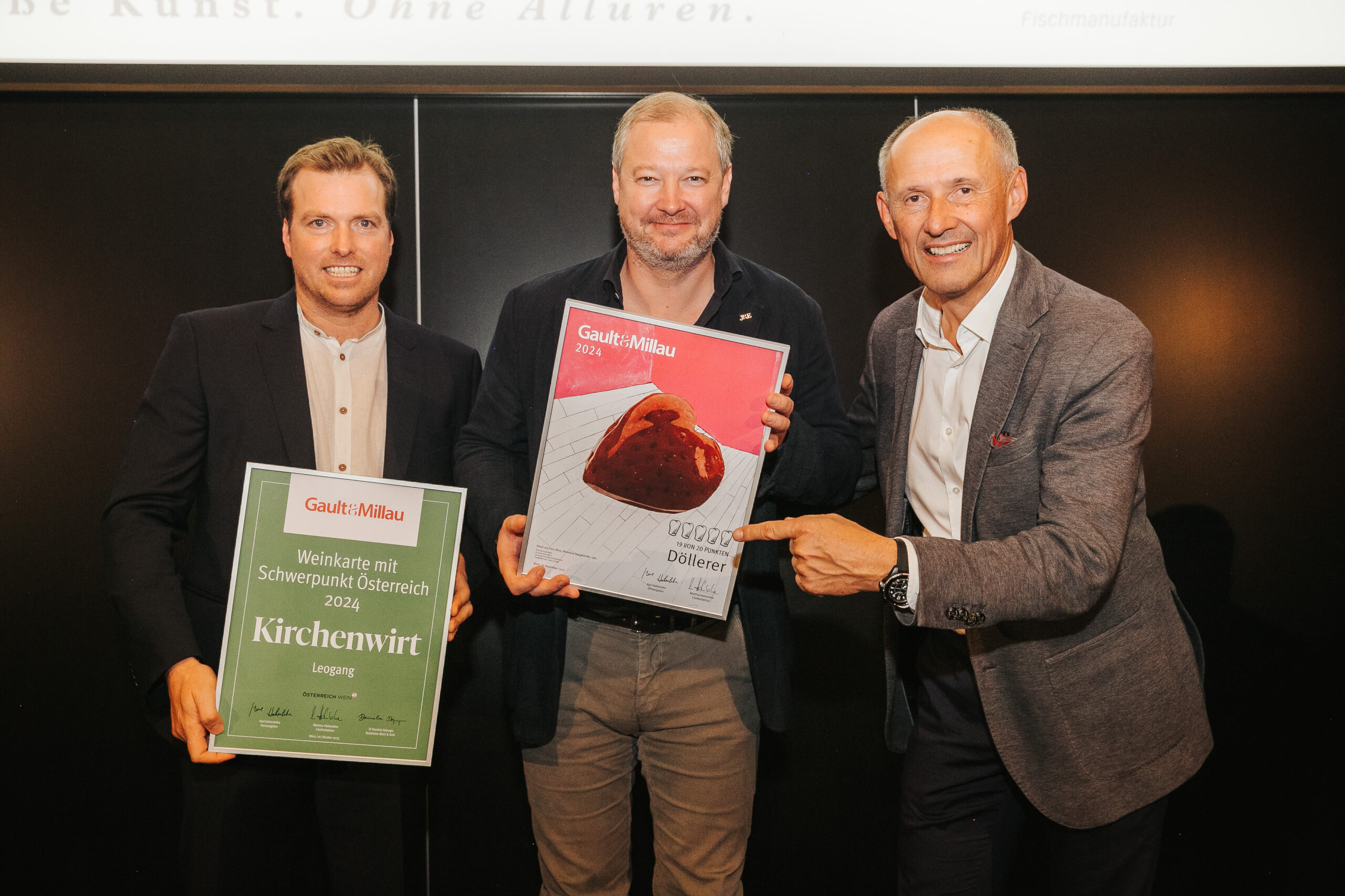 Drei Männer stehen lächelnd da, halten und zeigen auf die Gault&Millau 2024 Restaurant- und Kuchenpreise.