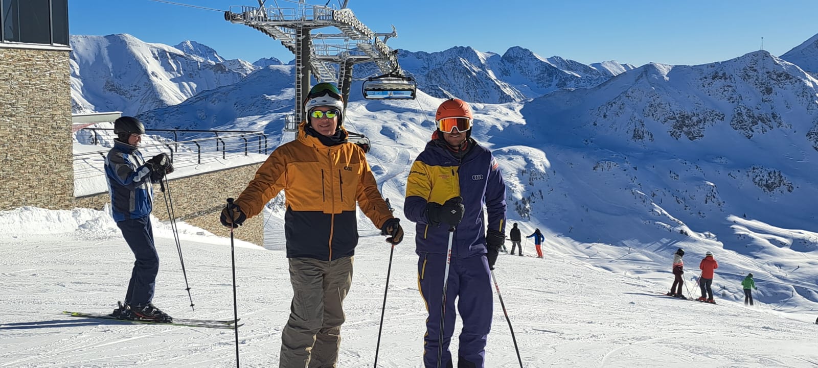 Zwei Skifahrer posieren an einem sonnigen Tag auf einer verschneiten Piste mit Bergen und einem Skilift im Hintergrund.
