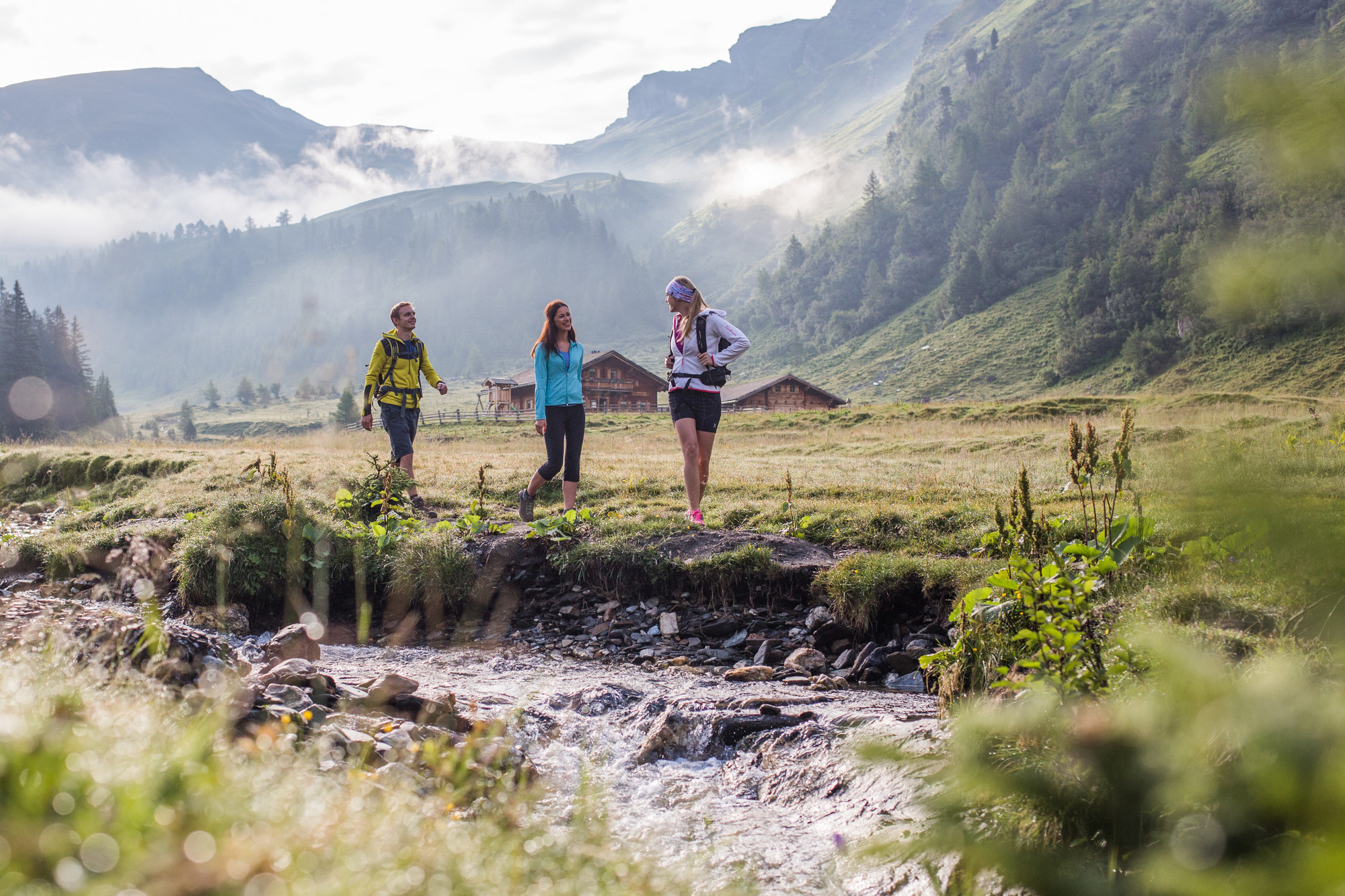 Drei Personen wandern entlang eines Baches in einer grasbewachsenen Berglandschaft mit Nebel und Hütten im Hintergrund.