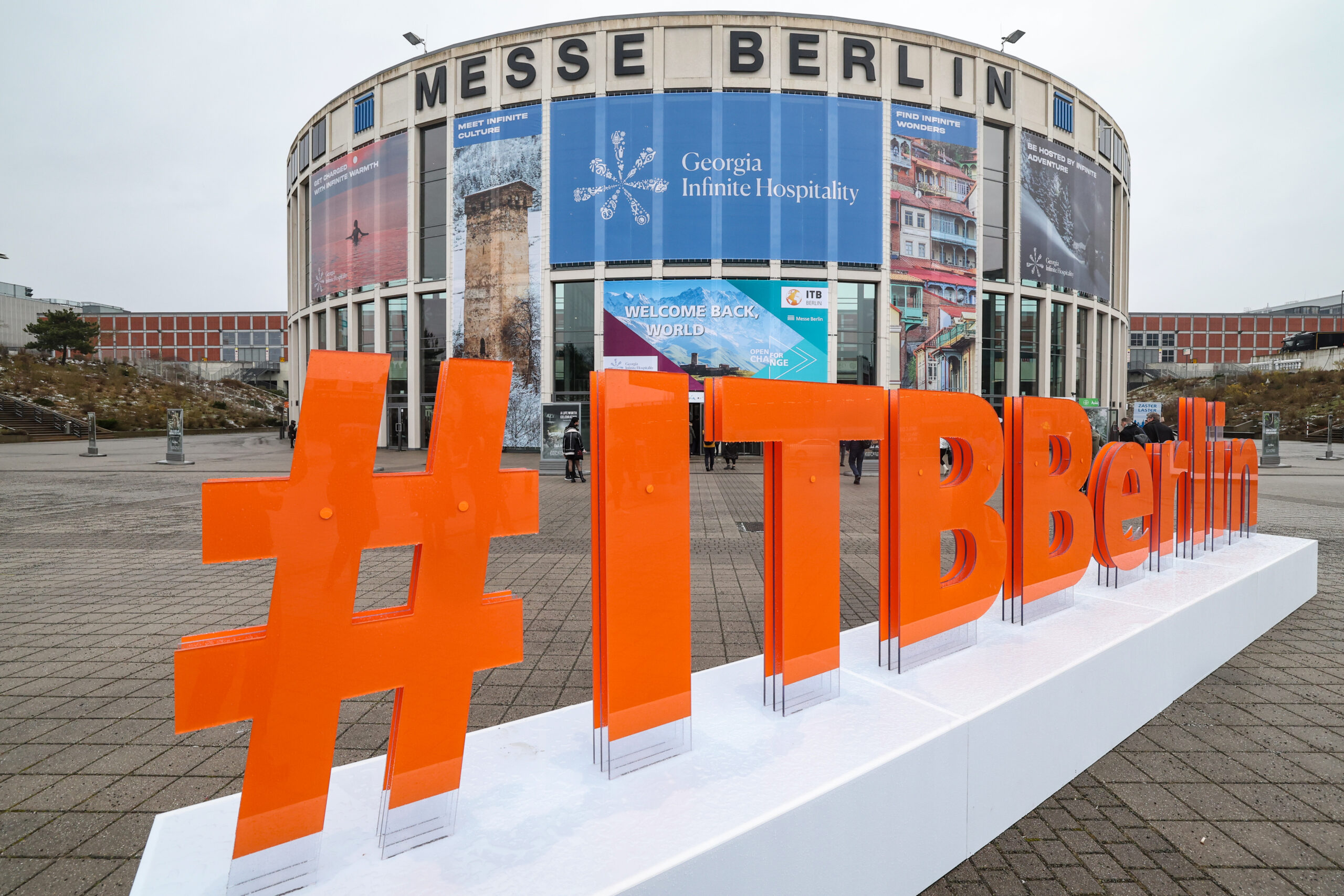 Großes orangefarbenes #ITBBerlin-Schild vor dem Gebäude der Messe Berlin mit darüber angebrachten Veranstaltungsbannern.