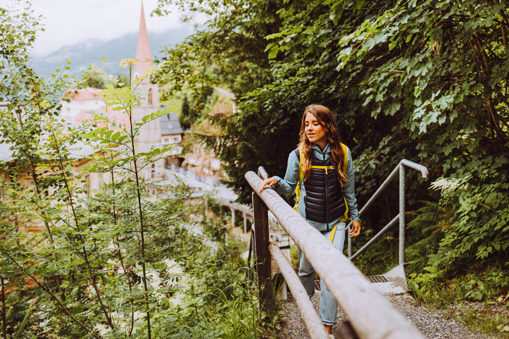Frau geht einen Waldweg hinauf, im Hintergrund sind ein Dorf und ein Kirchturm zu sehen.