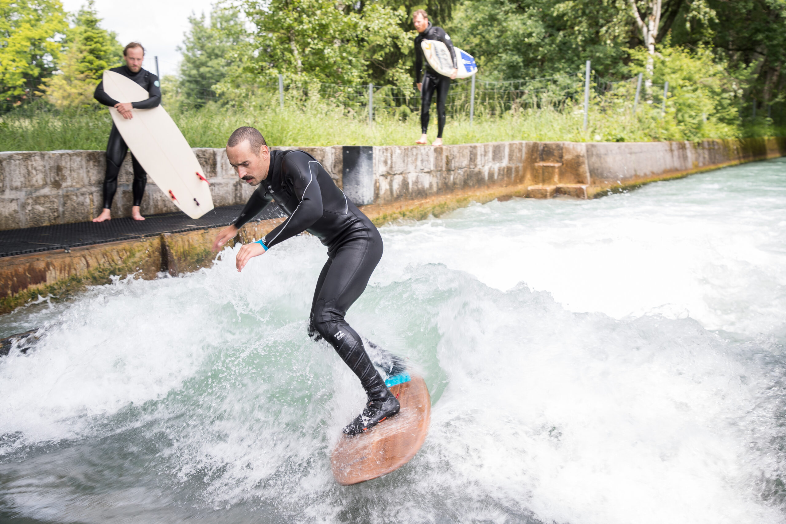 Ein Mann surft auf einer Flusswelle, während zwei Personen mit Surfbrettern vom Betonufer aus zusehen.