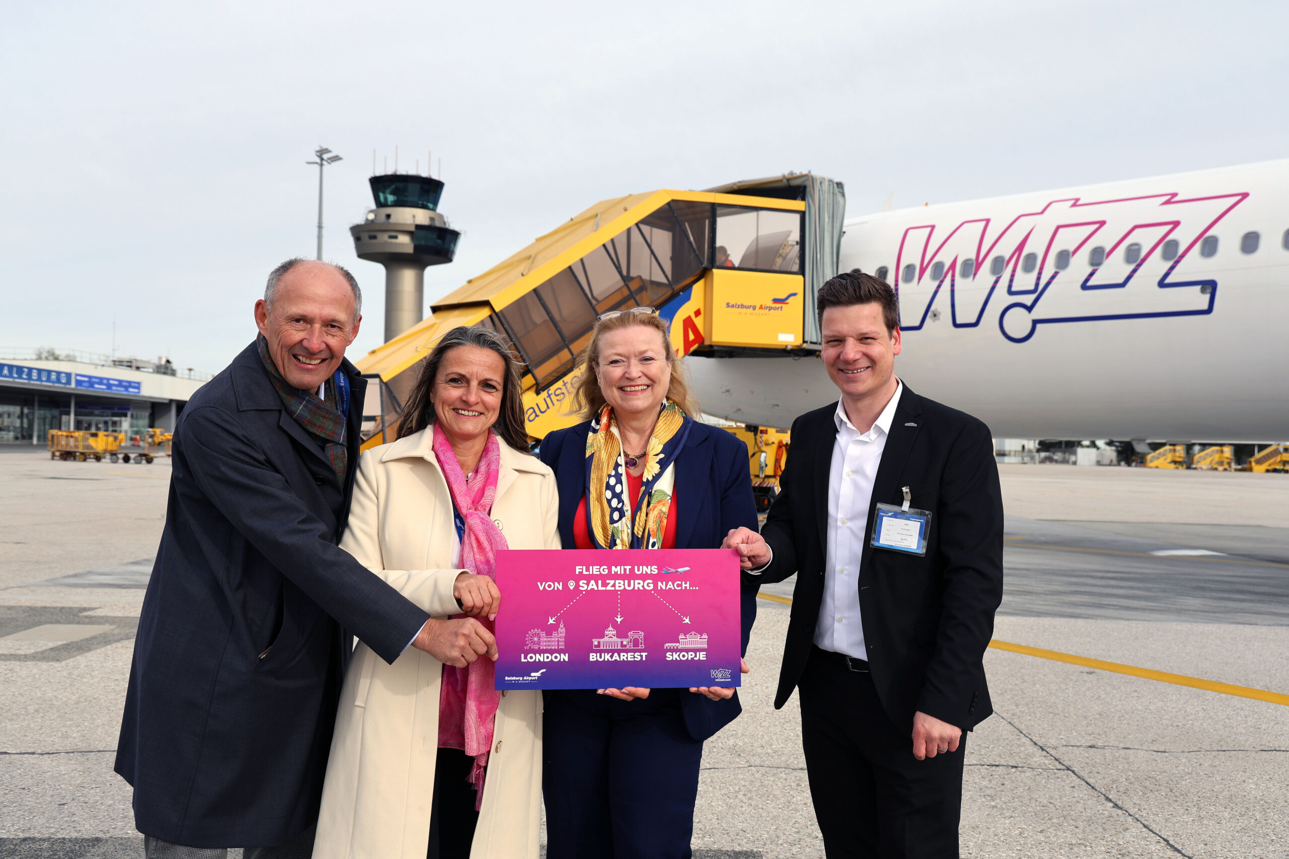 Vier Personen stehen lächelnd vor einem Flugzeug von Wizz Air und halten ein leuchtend rosa Zielschild in der Hand.