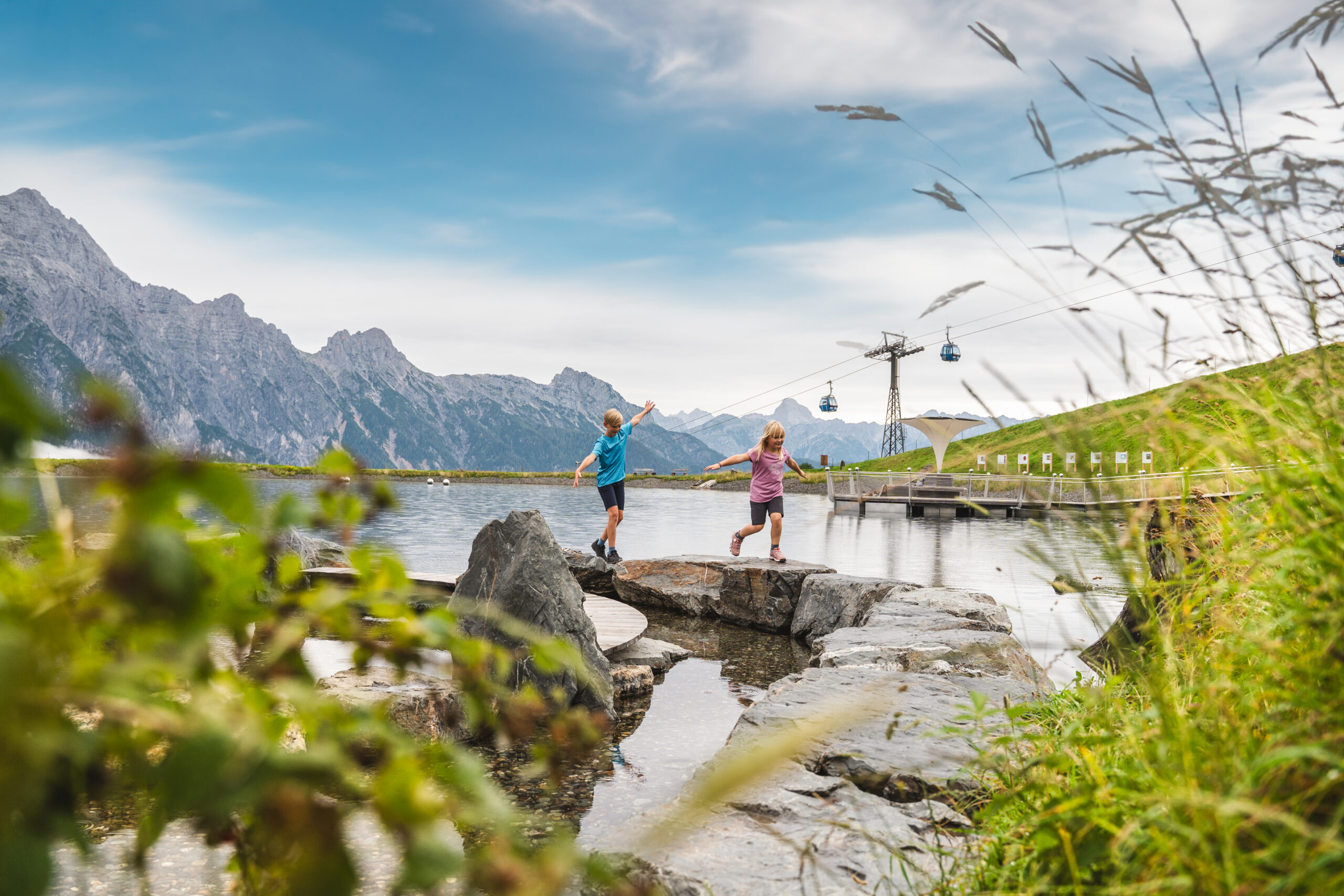 Zwei Kinder spielen auf Felsen neben einem Bergsee, mit Seilbahnen und Bergen im Hintergrund.