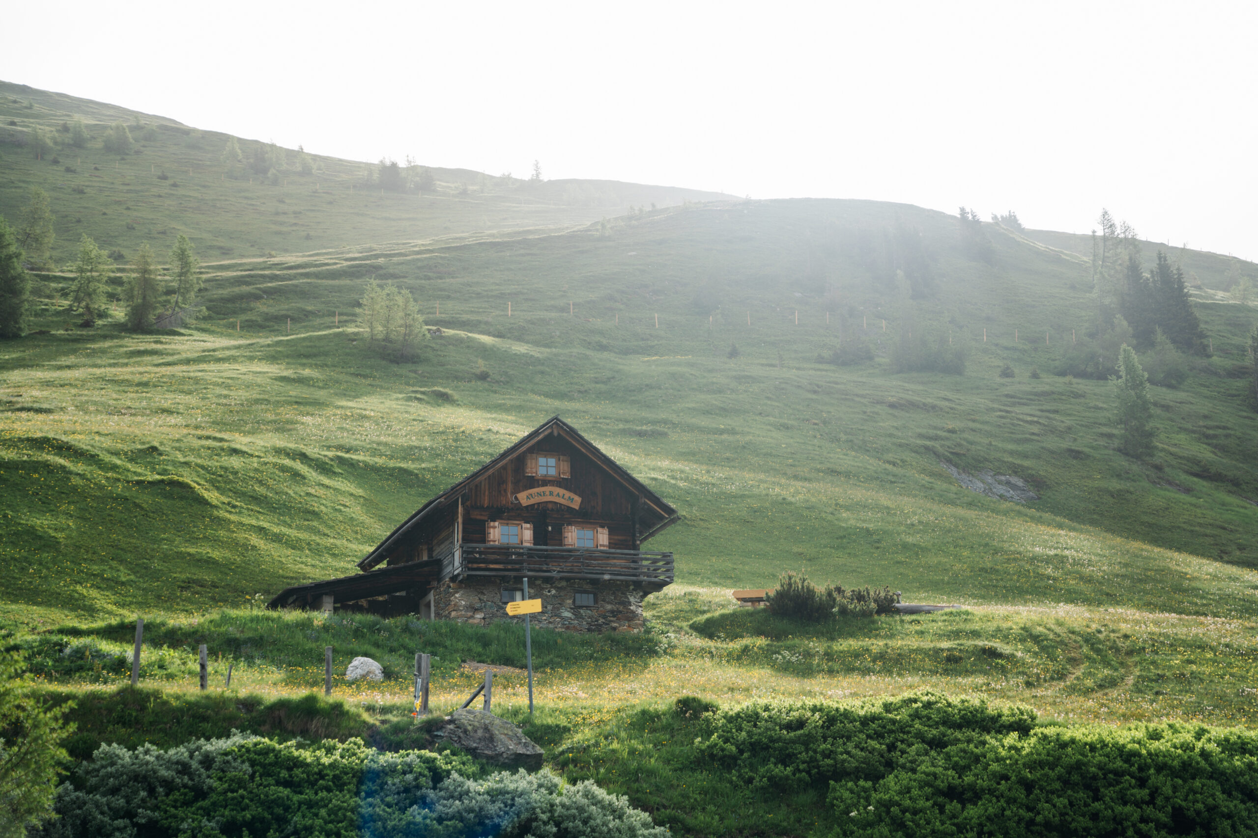Eine Holzhütte steht auf einem grasbewachsenen Hügel, umgeben von grünen Feldern und Bäumen unter einem strahlenden Himmel.