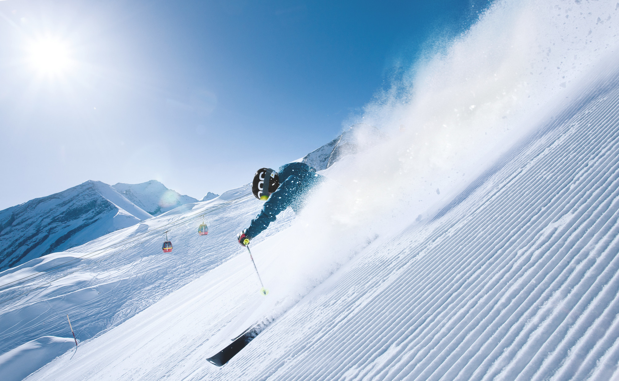 Ein Skifahrer rast an einem sonnigen Tag einen verschneiten Berghang hinunter und wirbelt Pulverschnee auf, während im Hintergrund Skilifte zu sehen sind.