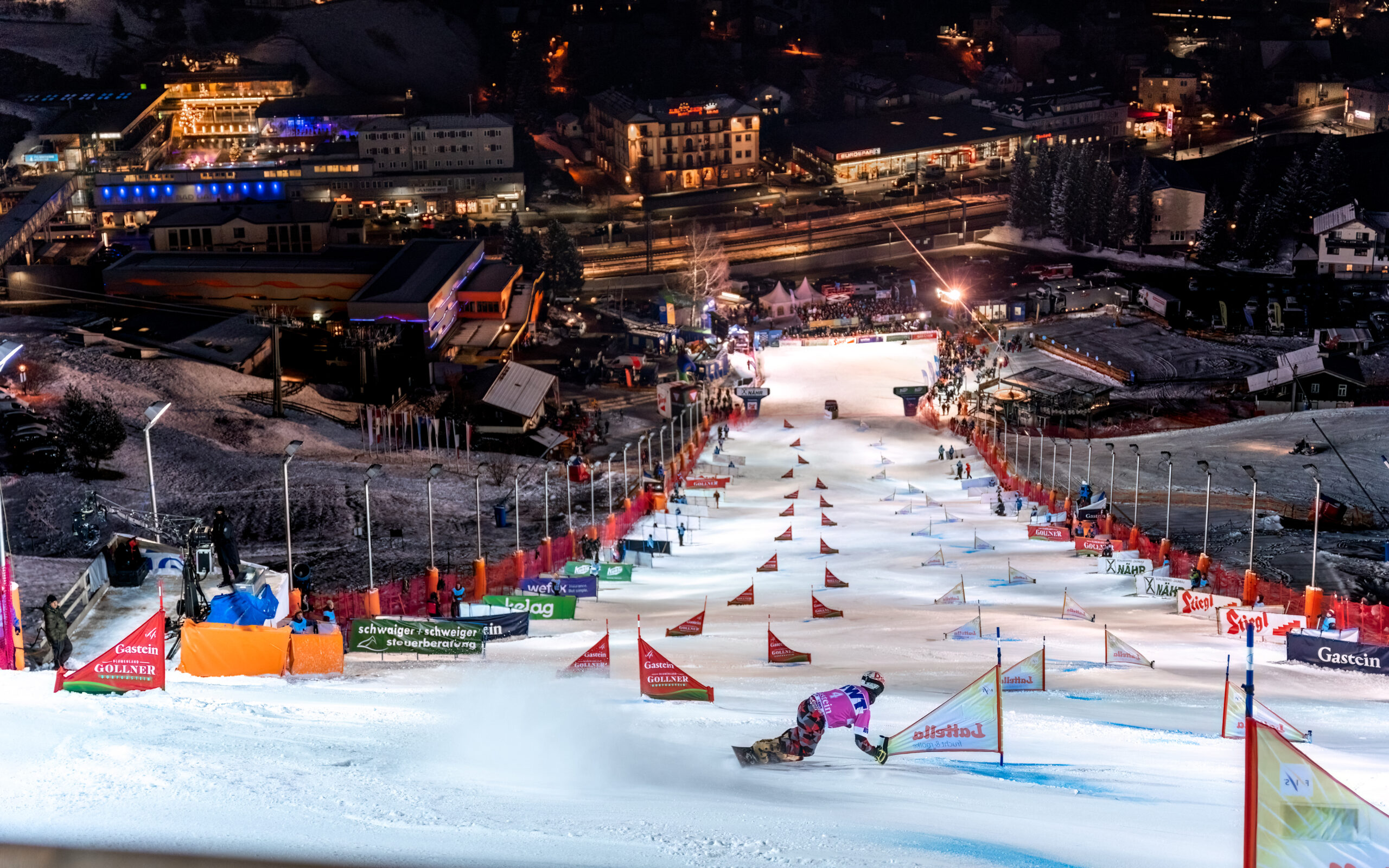 Ein Snowboarder fährt nachts eine Slalomstrecke hinunter, im Hintergrund sind Lichter und Gebäude zu sehen.