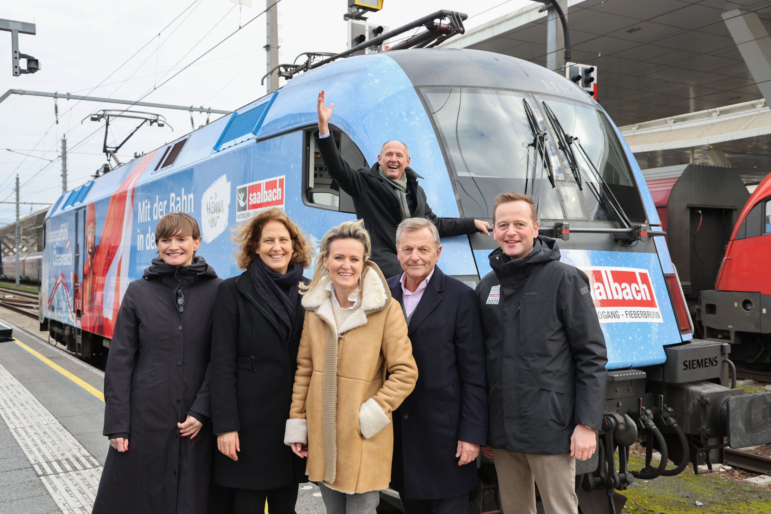 Sechs Personen posieren und lächeln vor einem blauen Zug; eine Person winkt aus dem Führerstand des Zuges.