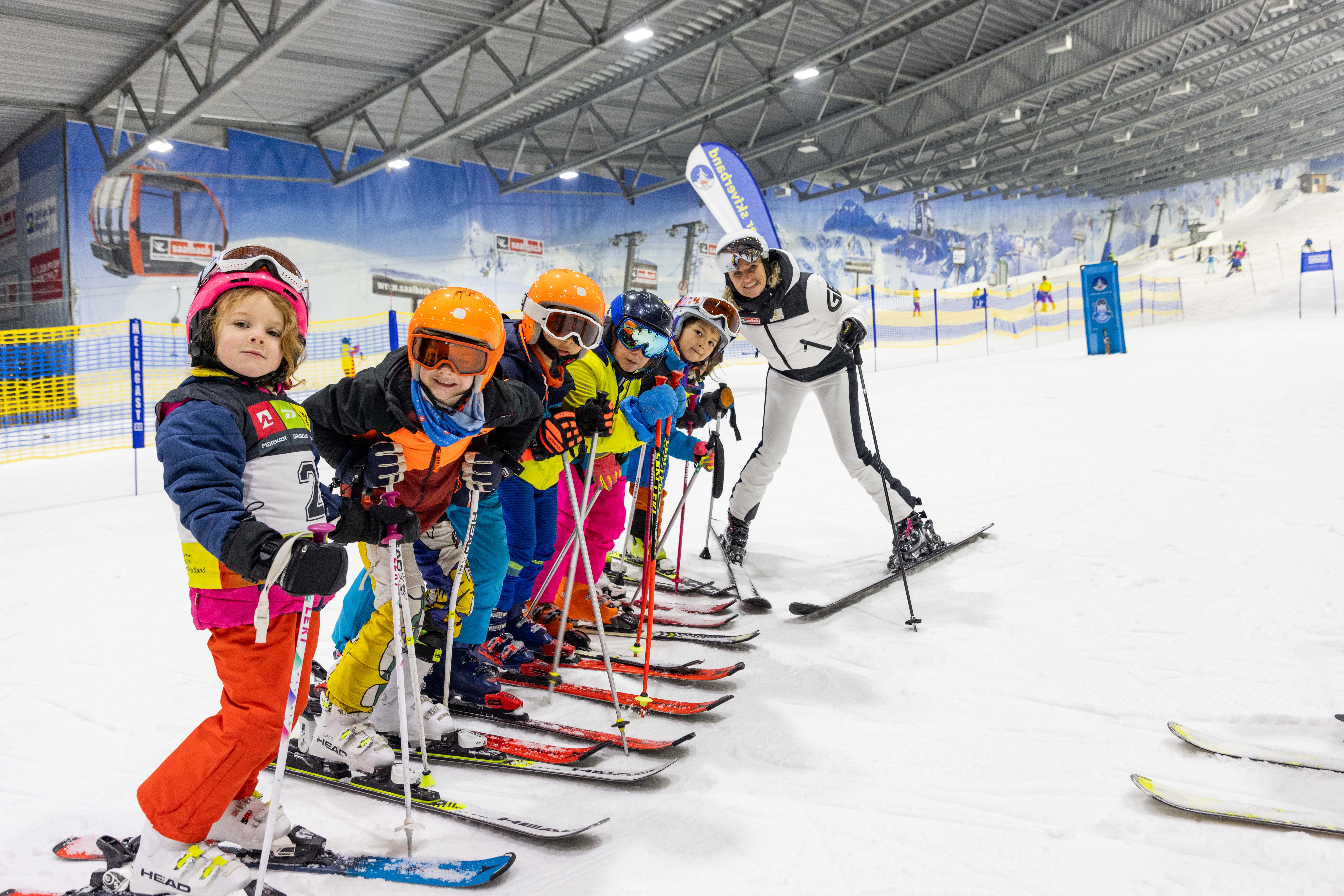 Ein Skilehrer posiert mit einer Gruppe von Kindern in farbenfroher Ausrüstung auf einer Indoor-Skipiste.