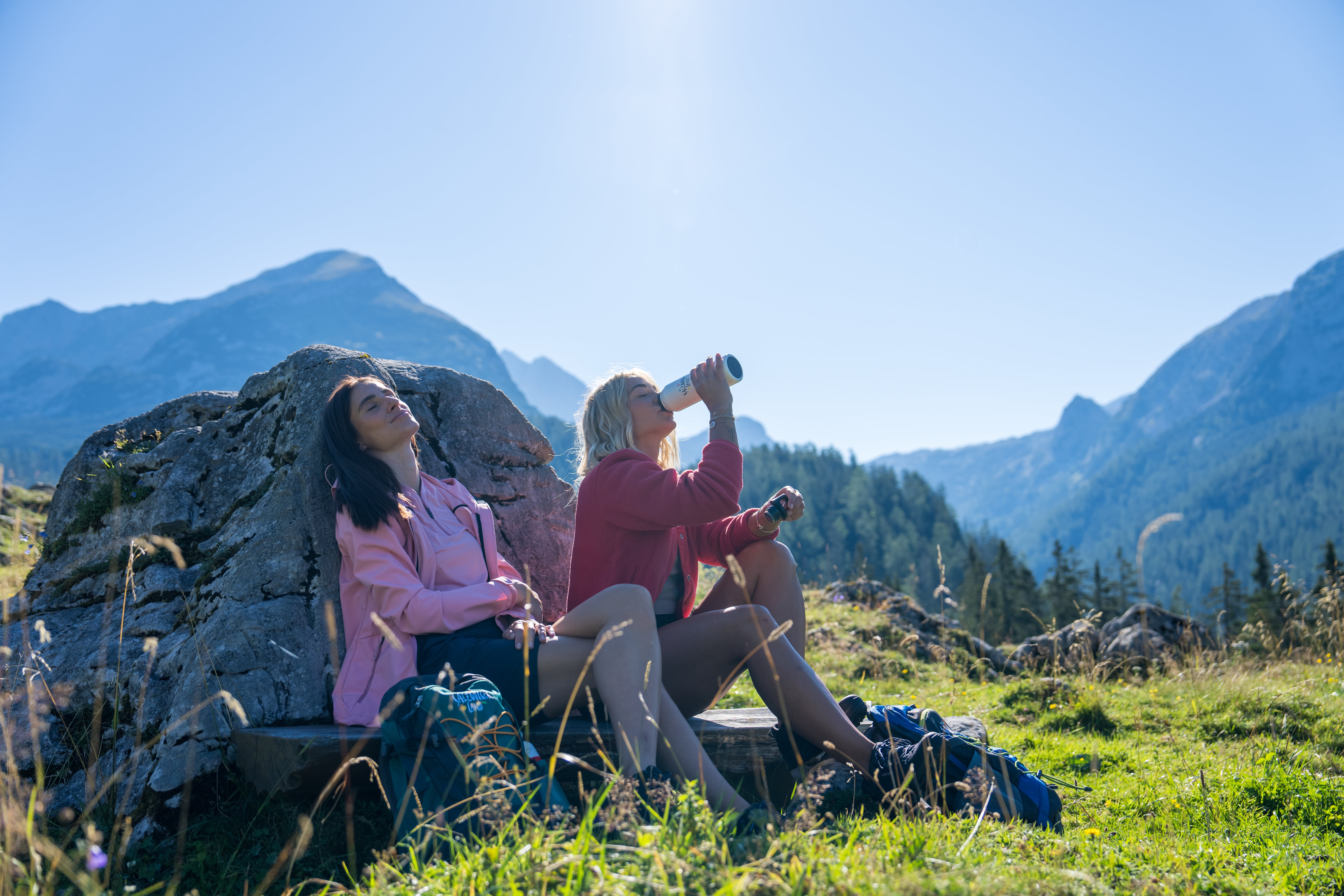 Zwei Frauen entspannen sich an einem Felsen in den Bergen; die eine trinkt aus einer Flasche, die andere genießt die Sonne.