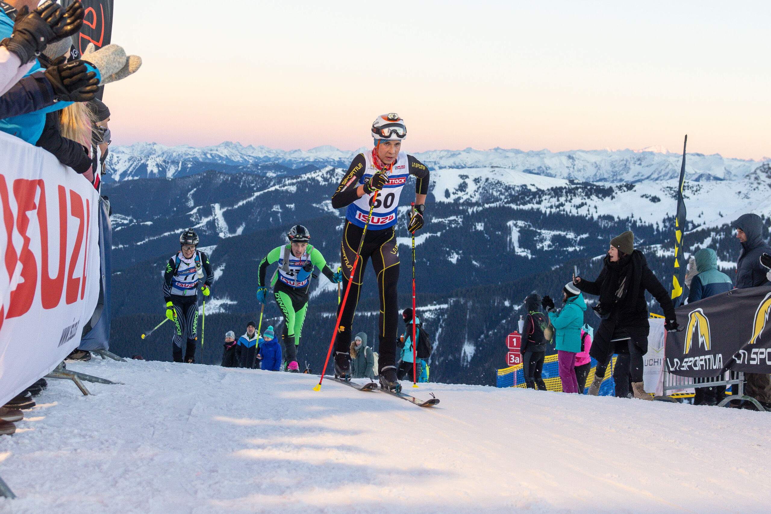 Skifahrer, die auf einem verschneiten Berg bergauf fahren, mit Zuschauern und alpiner Landschaft im Hintergrund.
