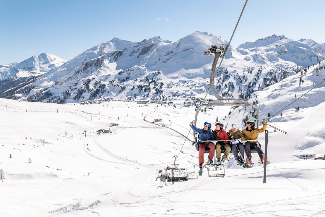 Vier Personen fahren an einem sonnigen Tag mit einem Skilift über verschneite Berge, umgeben von einer Winterlandschaft.