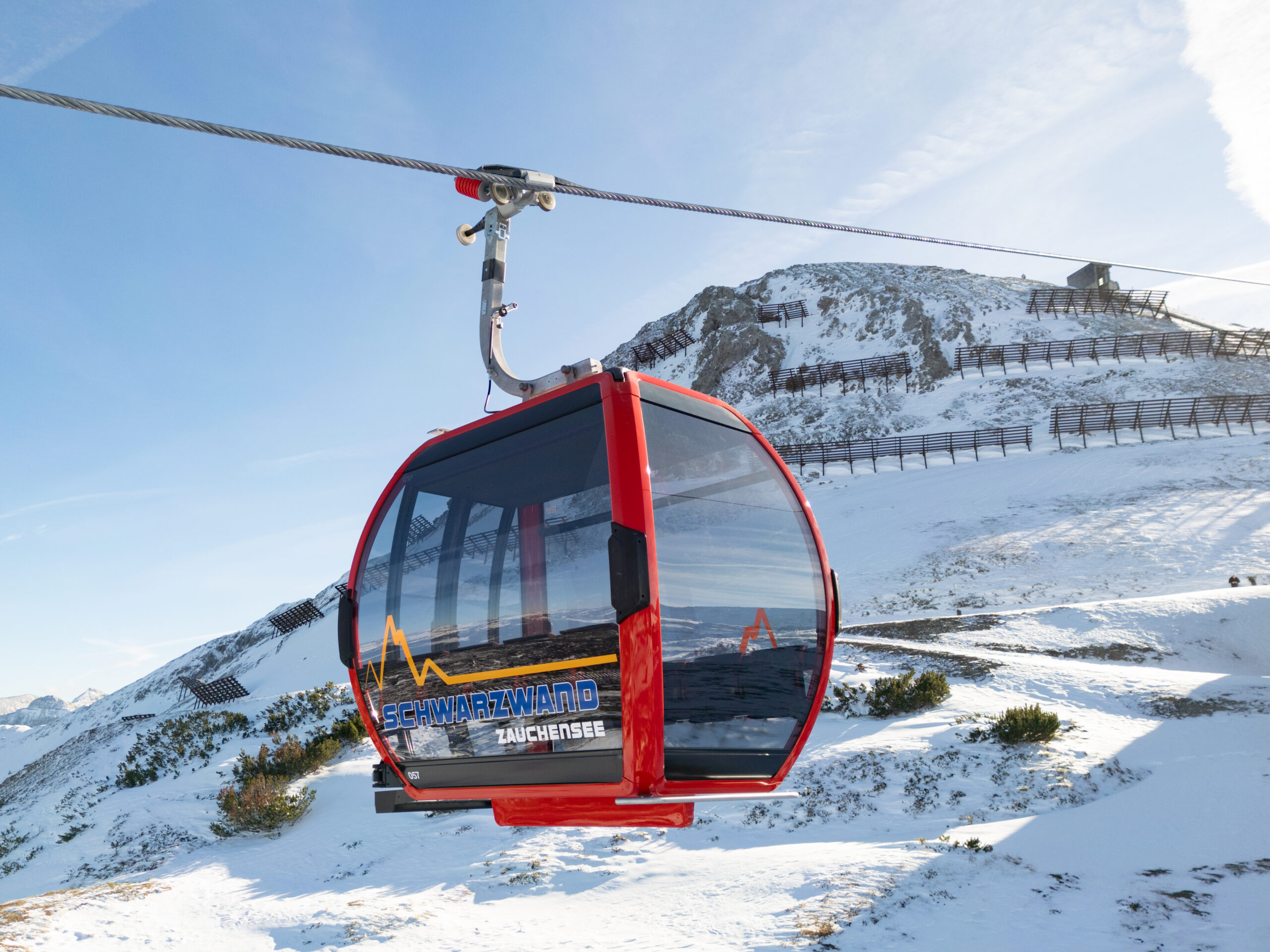 Rote Skigondel, die über einer verschneiten Berglandschaft mit blauem Himmel im Hintergrund schwebt.