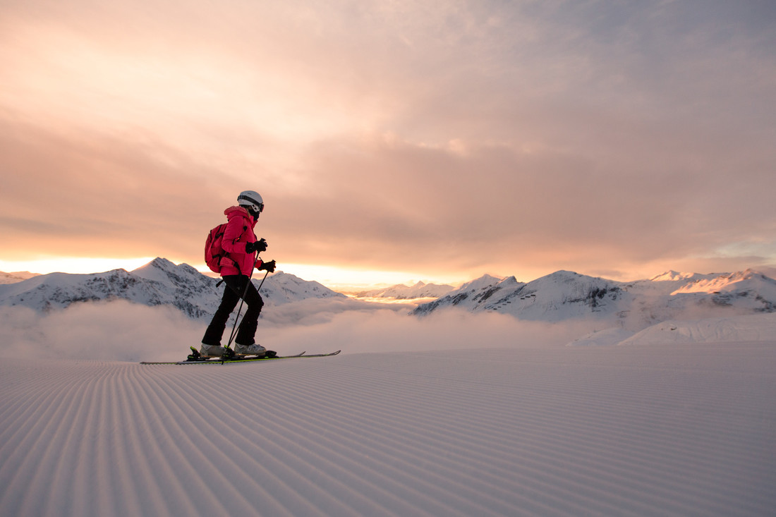 Person beim Skifahren bei Sonnenaufgang auf einer präparierten Piste mit schneebedeckten Bergen im Hintergrund.