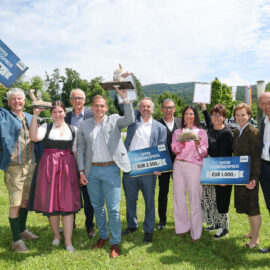 Eine Gruppe lächelnder Menschen im Freien, die Preisschilder und Trophäen halten und an einem sonnigen Tag feiern.
