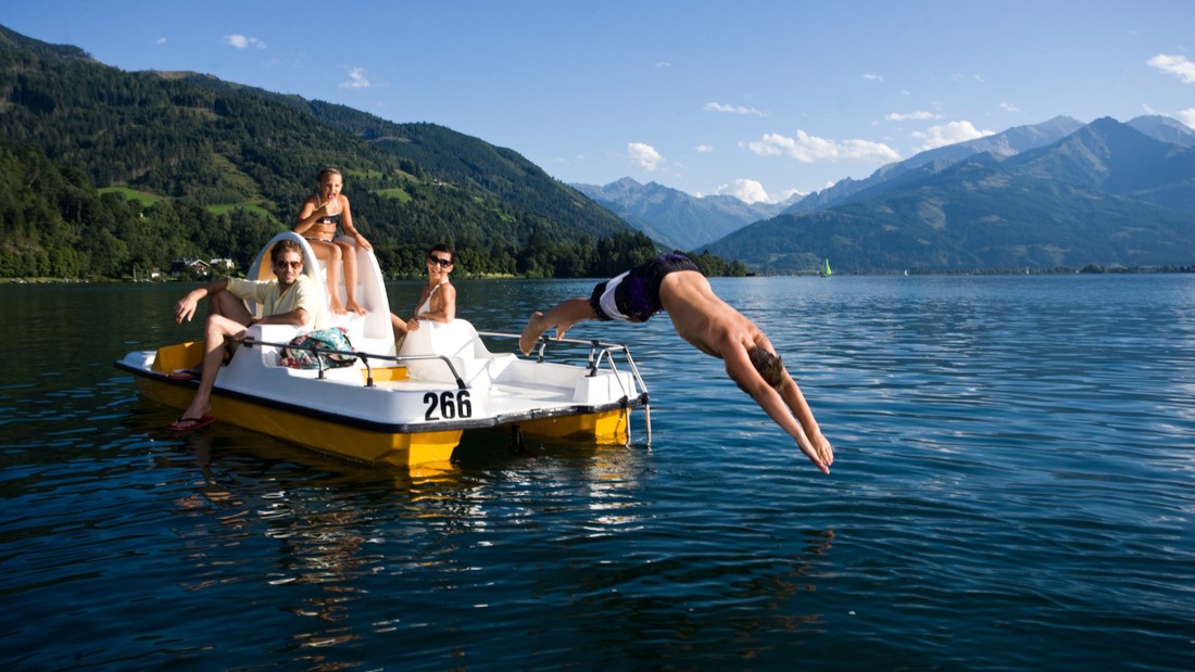 Vier Personen sitzen auf einem Tretboot auf einem See, ein Mann springt ins Wasser, im Hintergrund sind Berge zu sehen.