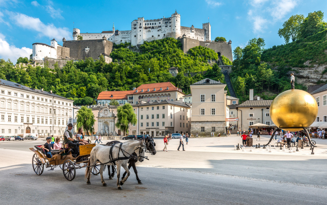 Eine Pferdekutsche auf einem sonnigen Stadtplatz mit einer Festung auf einem Hügel und einer großen goldenen Kugelskulptur.