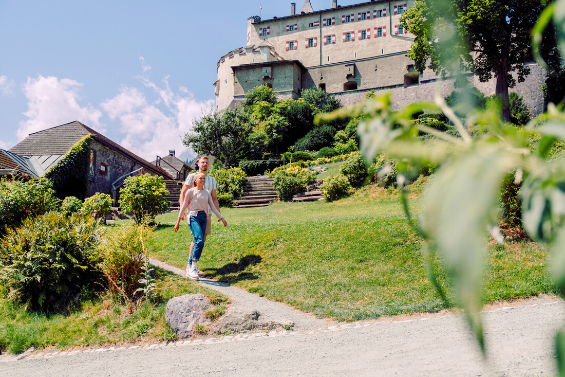 Ein Paar geht an einem sonnigen Tag einen Gartenweg hinauf in Richtung eines großen historischen Gebäudes.