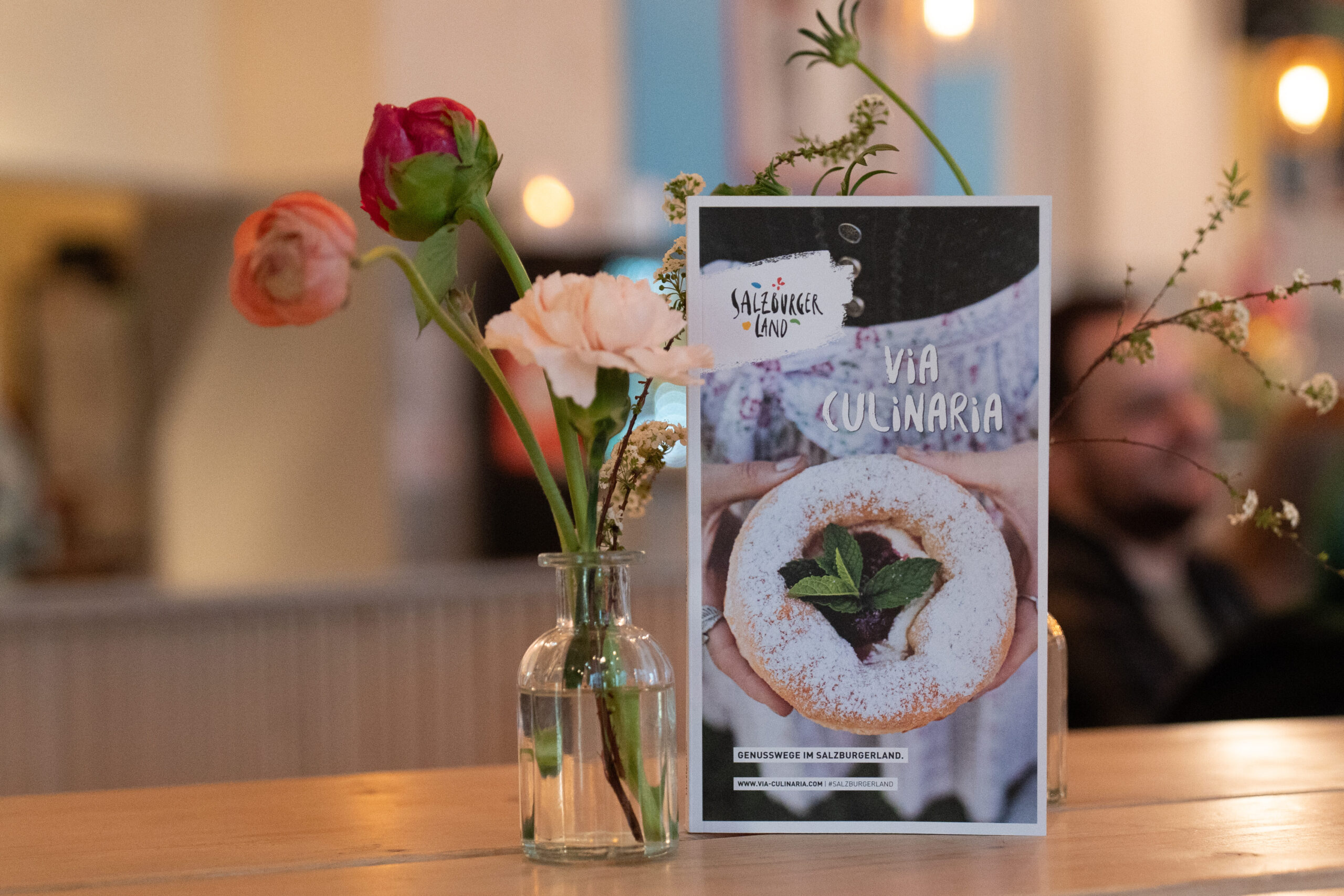 Eine kleine Blumenvase und eine Dessertkarte stehen auf einem Holztisch in einem gemütlichen, gedämpft beleuchteten Restaurant.