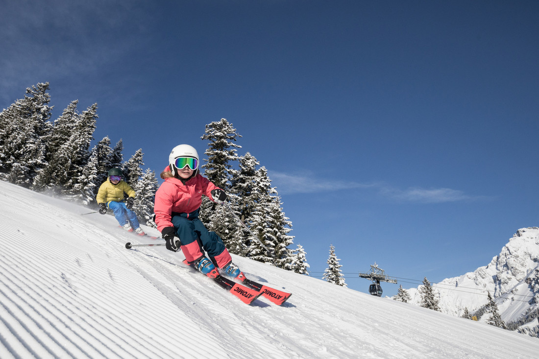 Zwei Personen fahren auf einer präparierten, verschneiten Piste mit Bäumen und Skilift im Hintergrund unter blauem Himmel Ski.