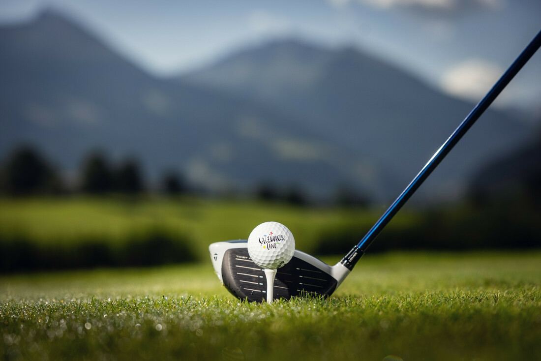 Ein Golfschläger, der so positioniert ist, dass er einen Golfball auf einem grasbewachsenen Platz mit Bergen im Hintergrund schlägt.