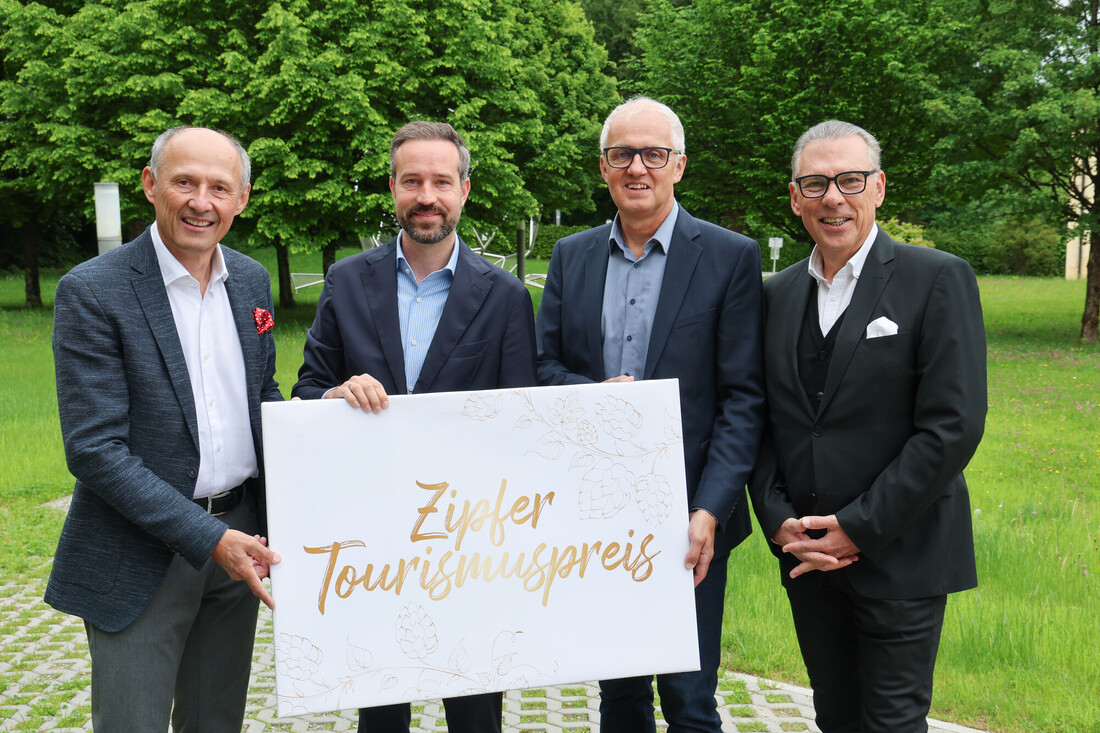 Vier Männer in Anzügen halten ein Schild mit der Aufschrift Zipfer Tourismuspreis im Freien mit grünen Bäumen im Hintergrund.